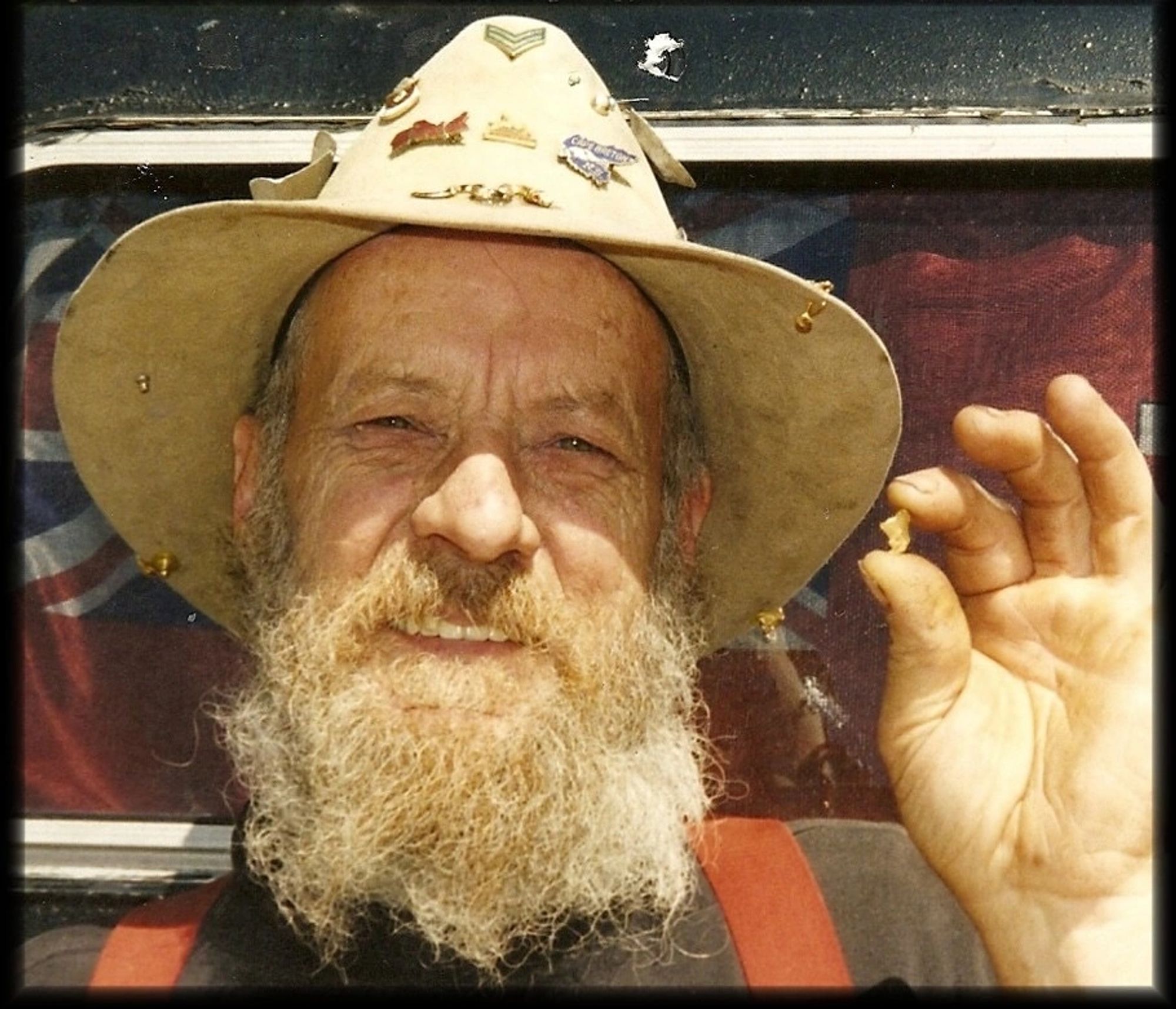 A bearded prospector in a hat holding up a small gold nugget